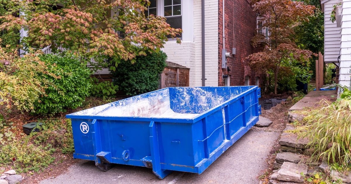 A blue roll-off dumpster with white staining on the inside parked in a residential driveway or alleyway between houses.