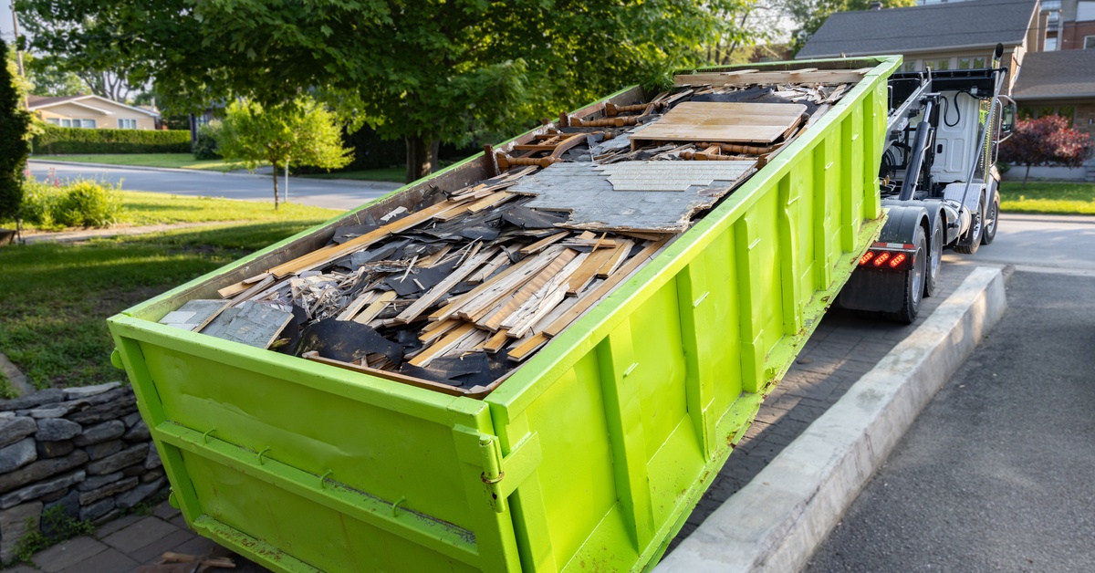 A lime green roll-off dumpster being delivered by a truck onto the driveway of a residential home.